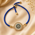 A blue braided cord bracelet with a gold-tone circular evil eye charm, displayed on a beige satin fabric background with dried flowers.