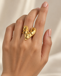 A close-up of a hand wearing a textured, sculptural gold ring shaped like a stylized heart or abstract form, set against a soft, light beige background.
