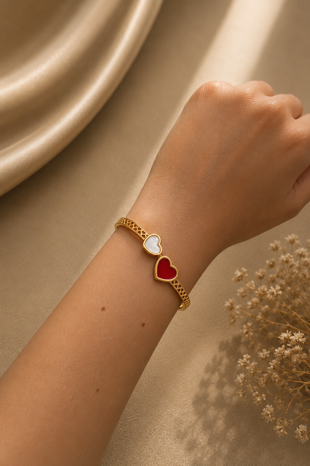 A gold chain bracelet worn on a person's wrist, featuring two dangling heart charms, one white and one red, set against a warm beige background with dried flowers.
